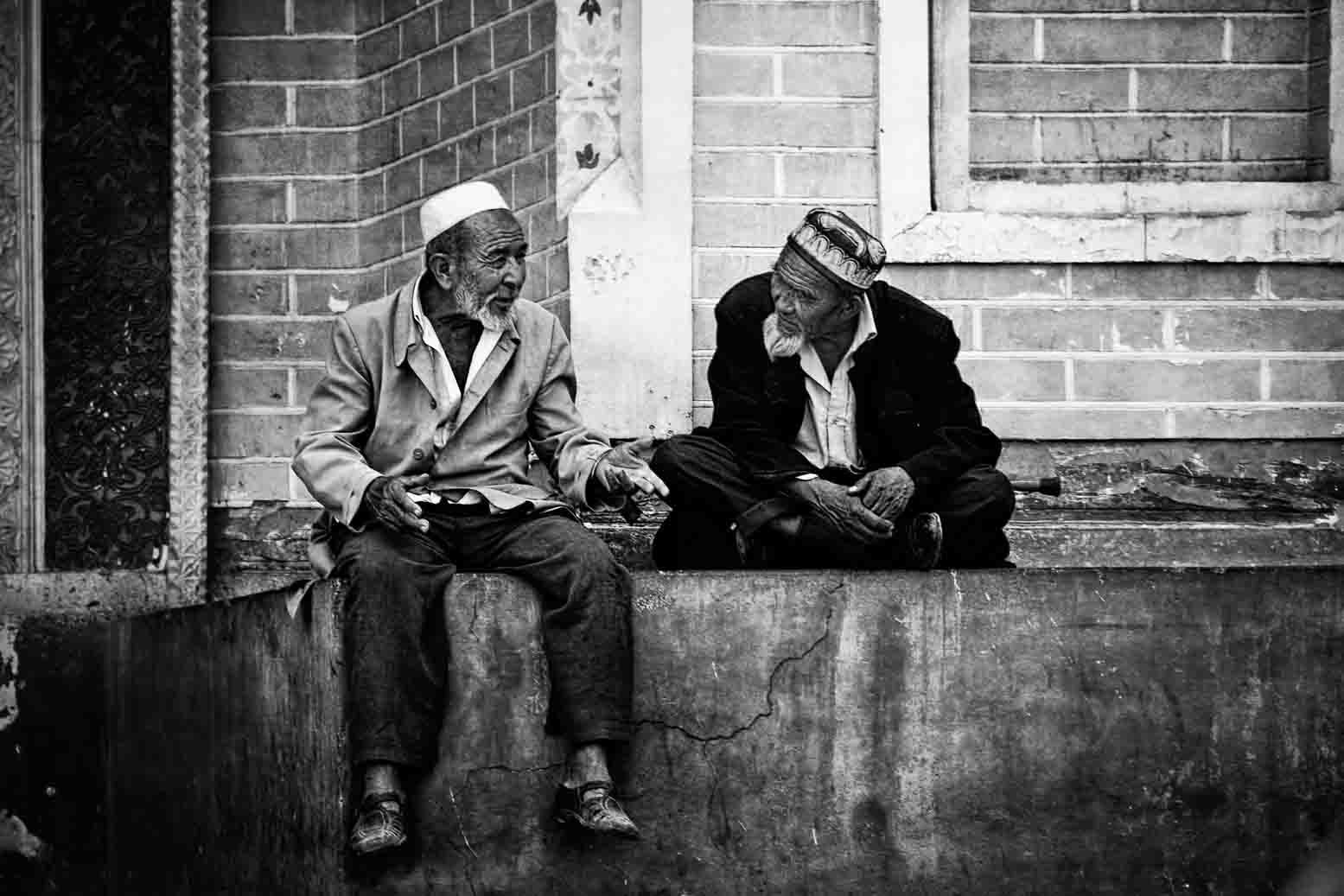 Two elders in doppa caps sitting on a low wall in conversation, in black and white