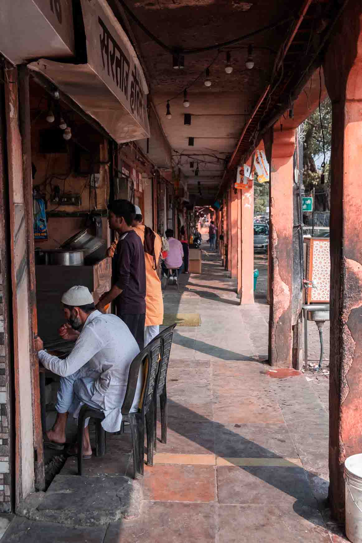 Men sitting outside a roadside stall under an orange colonnade