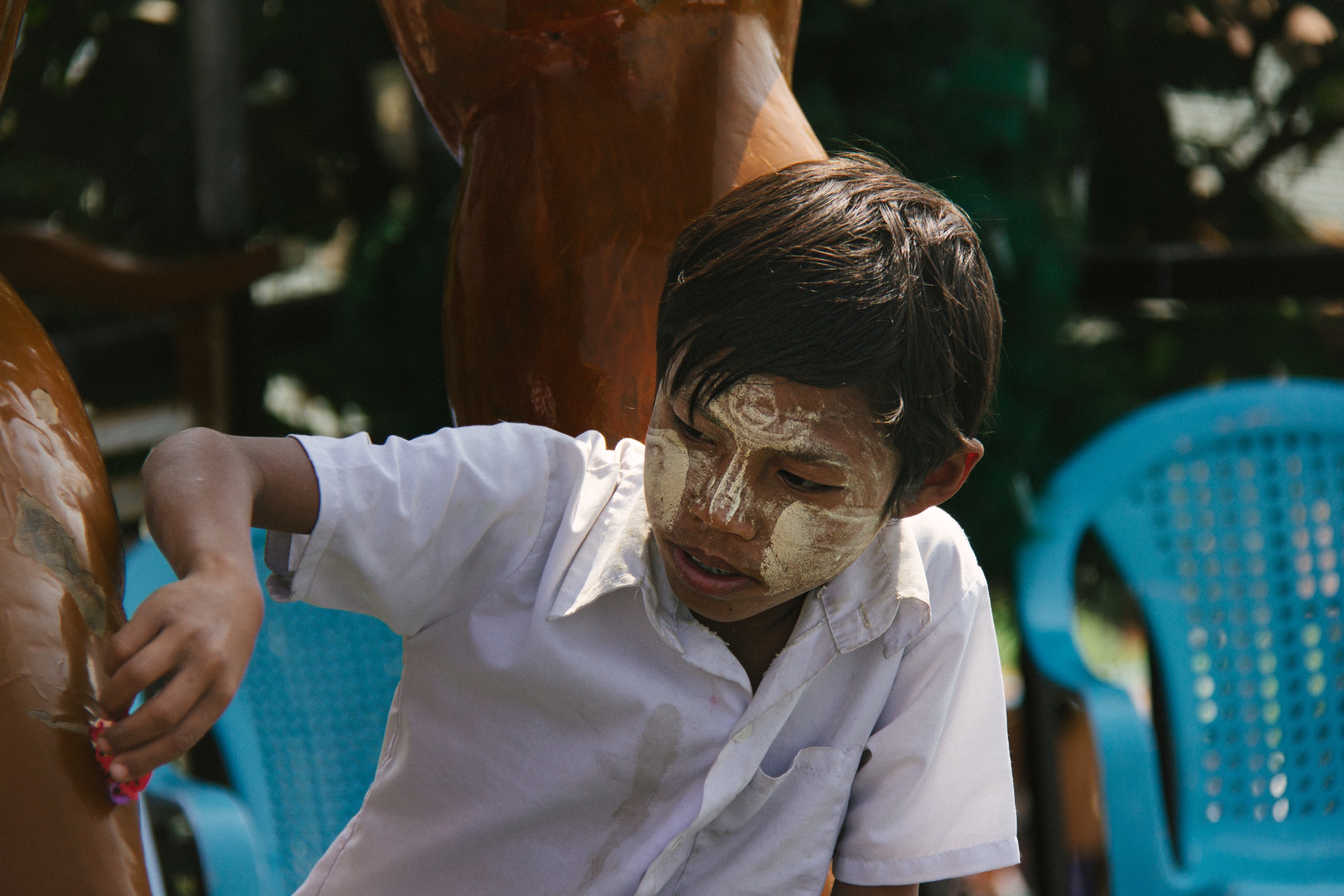 A boy in a white school shirt, cheeks painted with thanaka, glancing aside between blue plastic chairs
