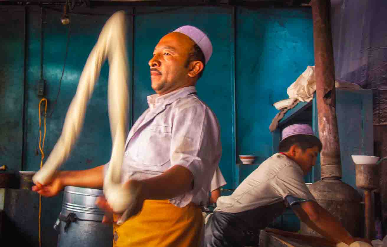 Two noodle-makers stretching ropes of dough in a turquoise-walled kitchen