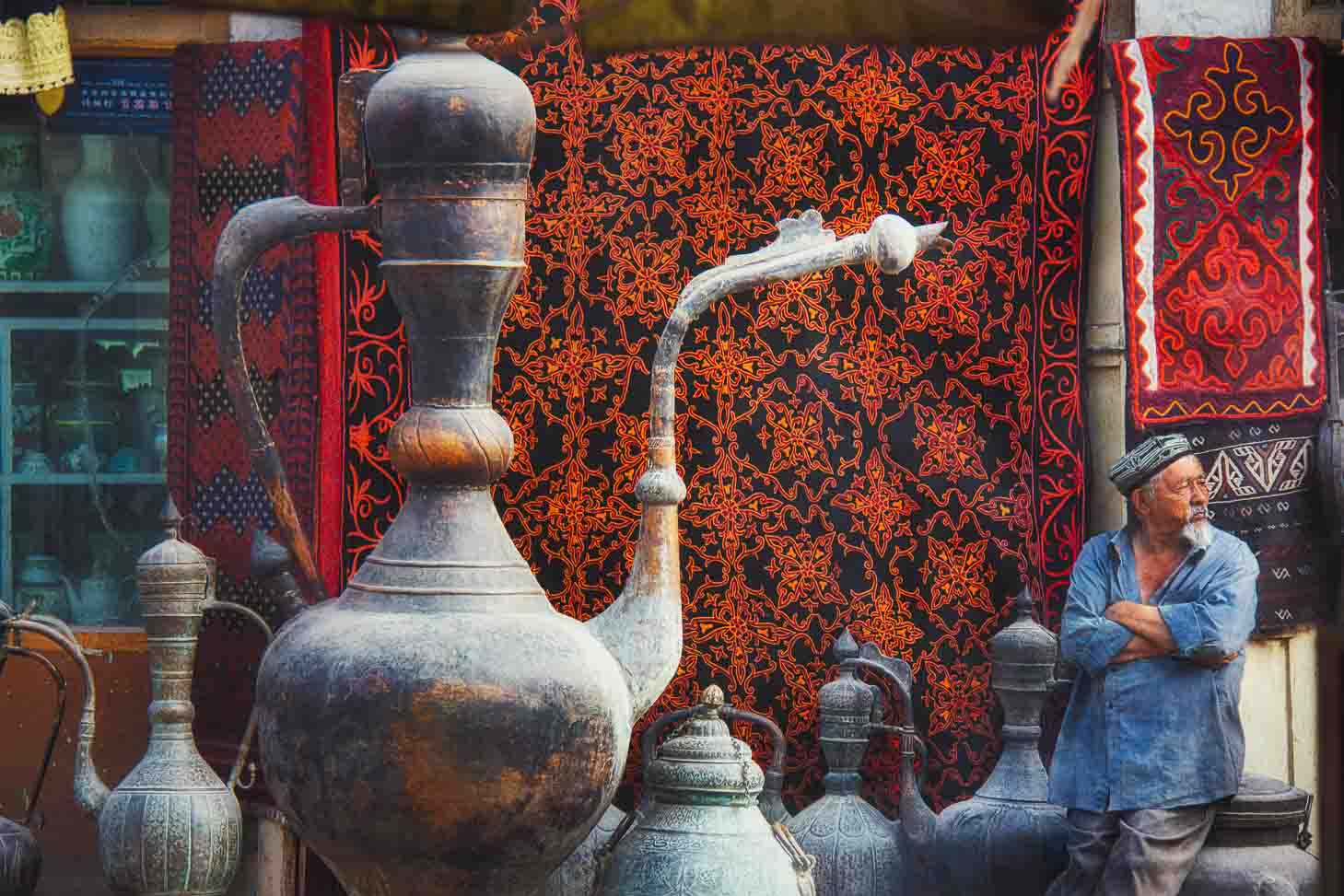 Elder vendor with arms folded amid hanging carpets and copper jugs