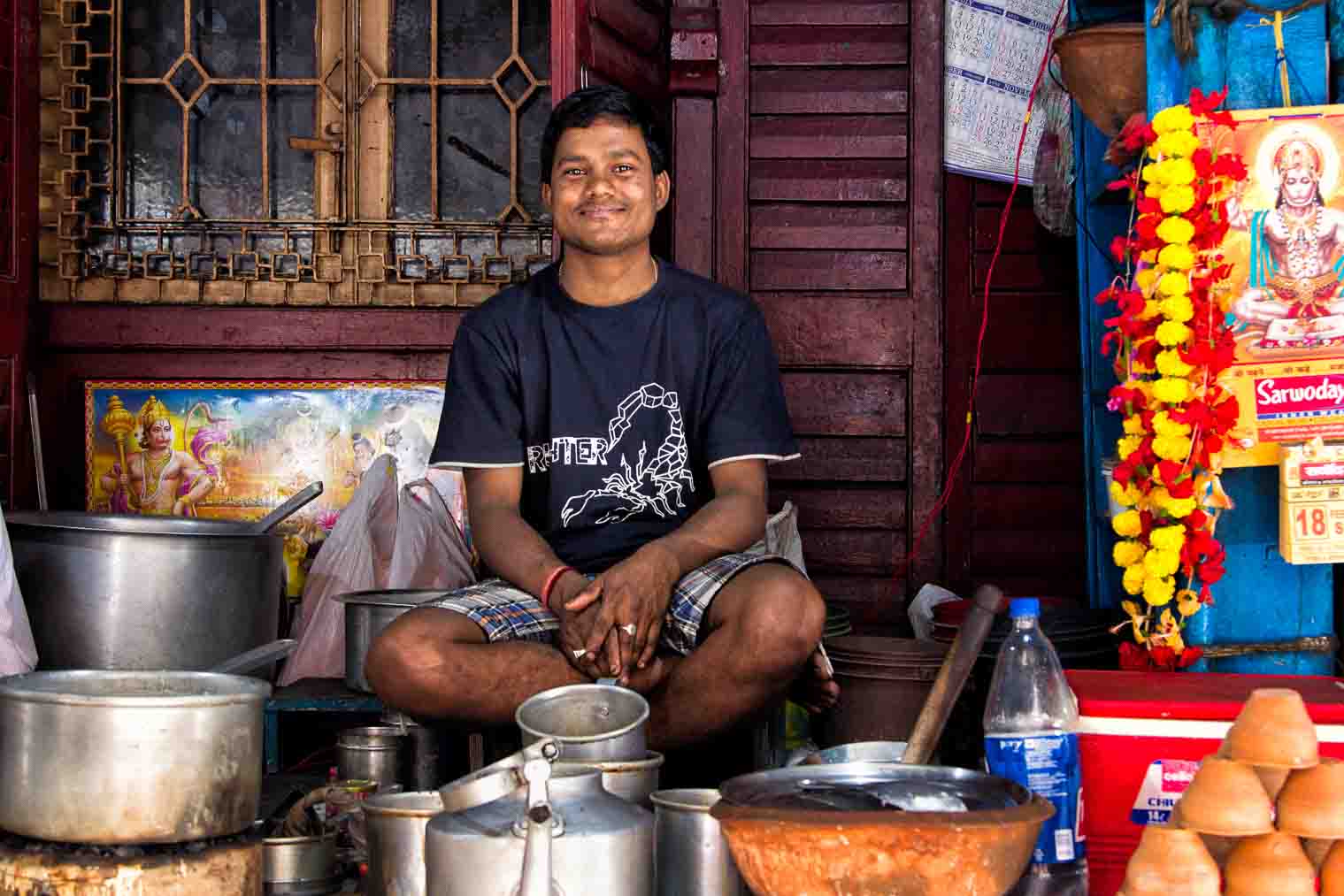 Chai-stall vendor seated cross-legged behind kettles, garlands and a Hanuman poster behind