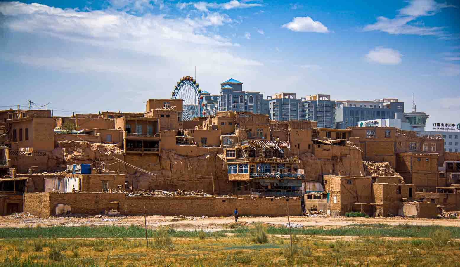 Mud-brick old town in mid-demolition, high-rise towers and a ferris wheel rising behind